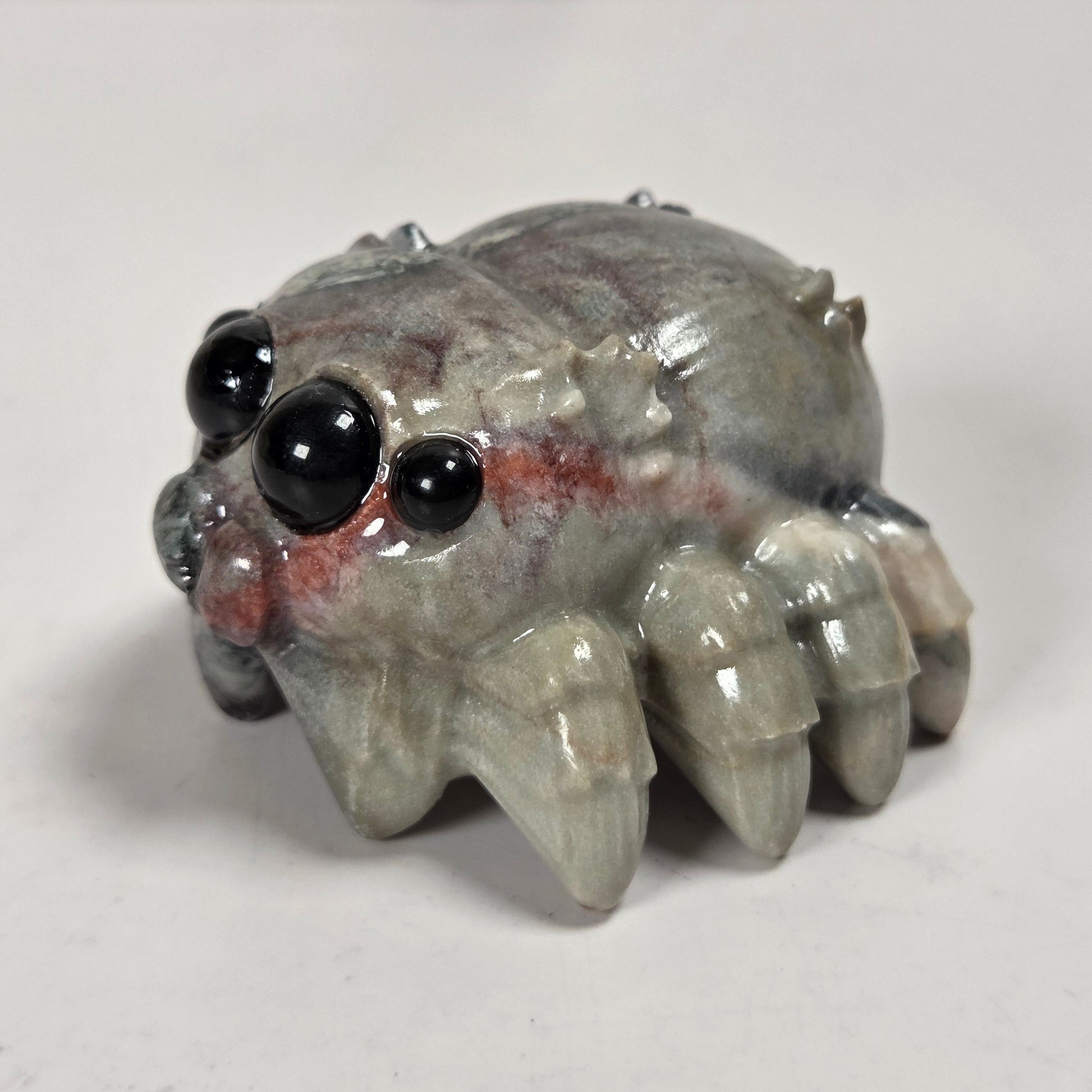 A small spider figurine with glossy black eyes and a gray body with subtle red markings, photographed on a plain light background.