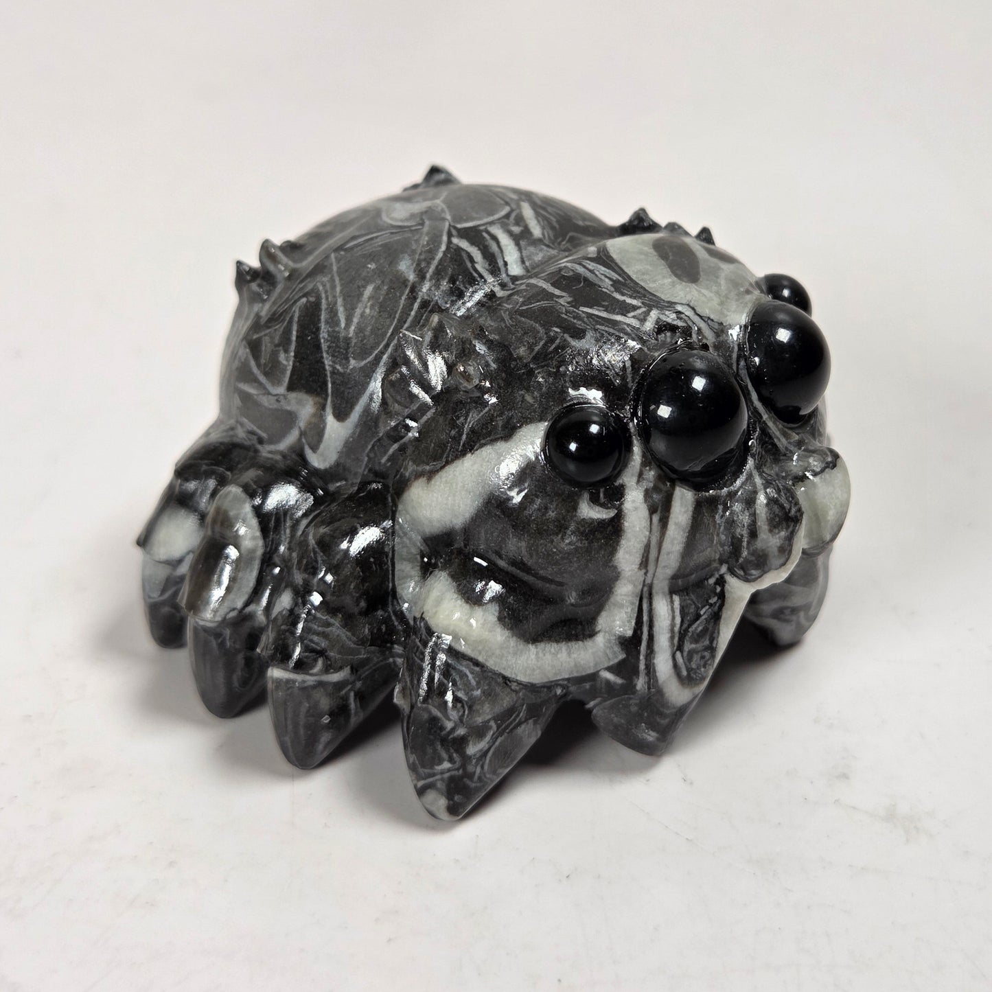 A small carved figurine of a spider made from polished black and white marbled stone, featuring glossy black eyes, set against a plain light gray background.
