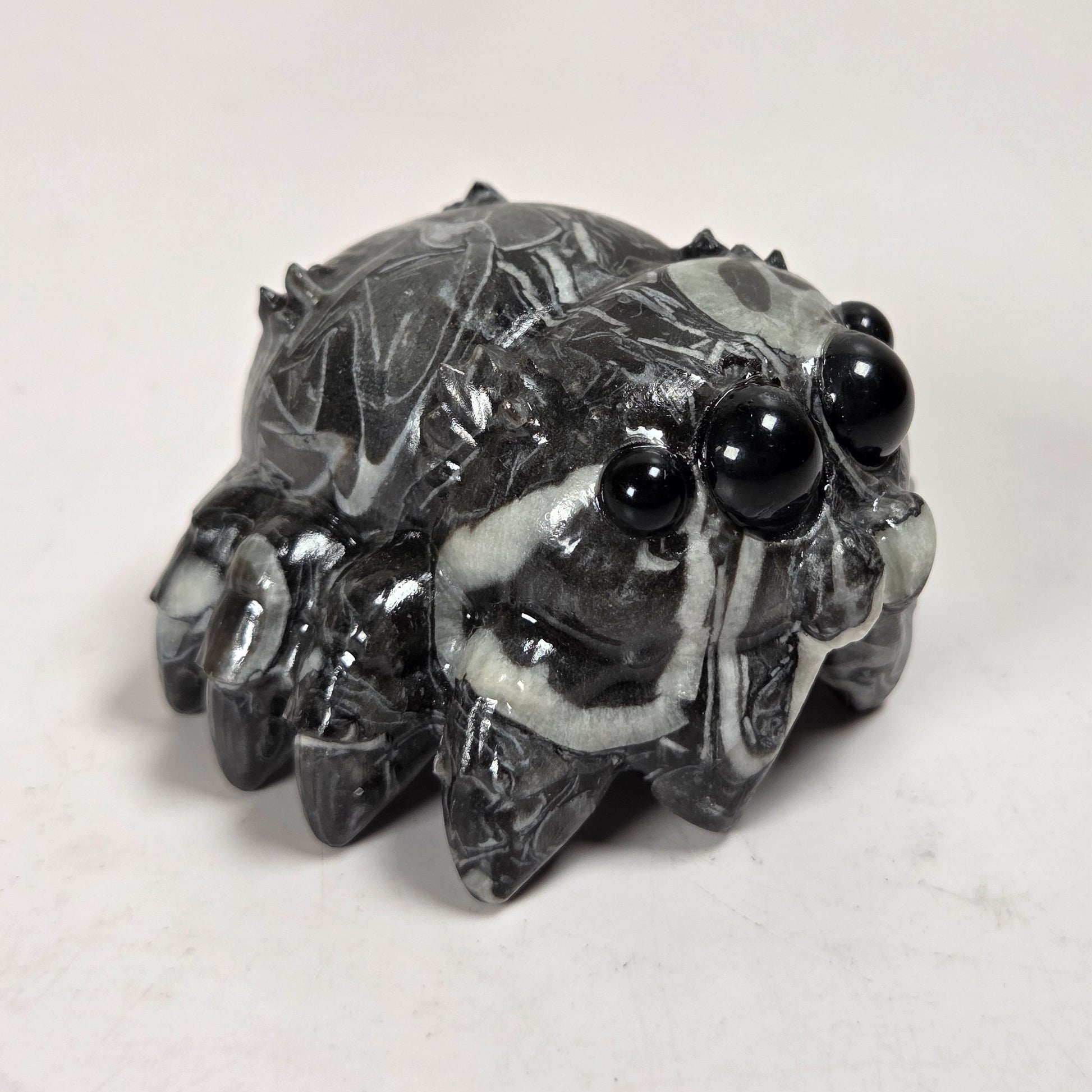 A small carved figurine of a spider made from polished black and white marbled stone, featuring glossy black eyes, set against a plain light gray background.