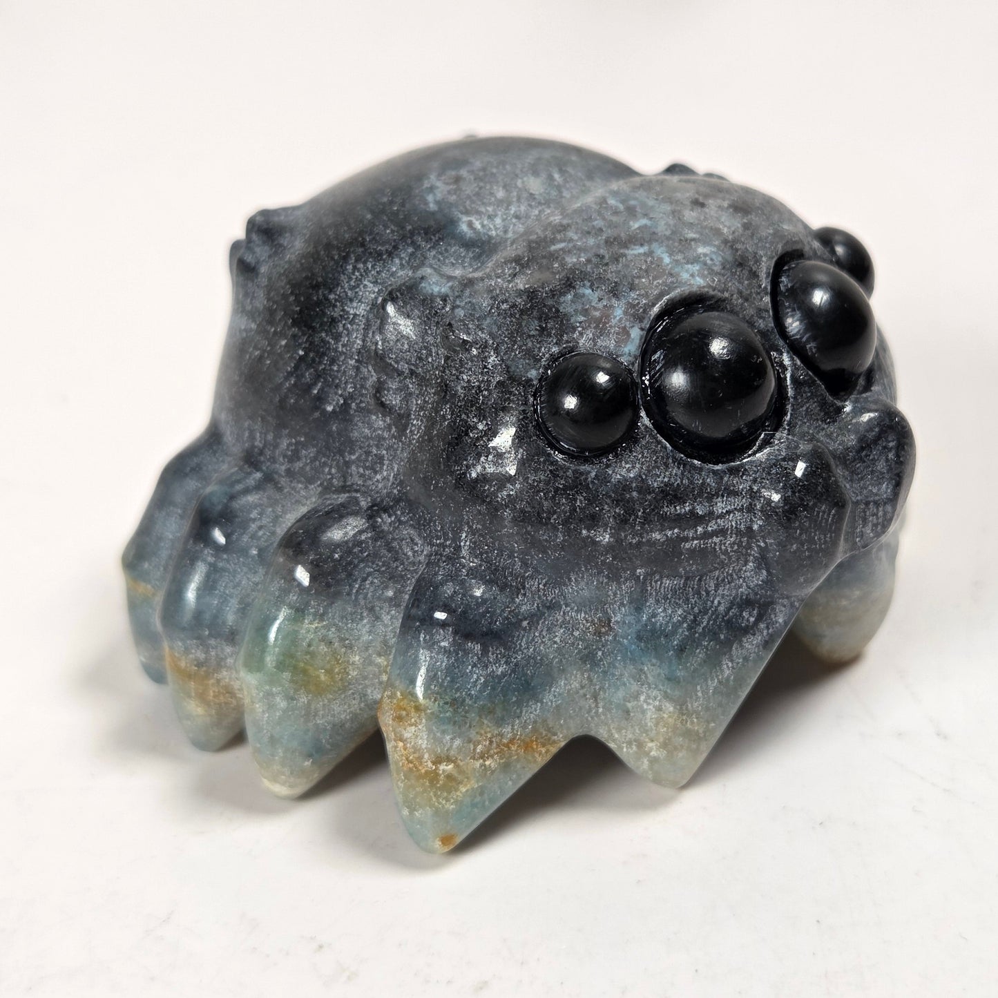 A small carved stone creature figurine with a textured, weathered finish in shades of gray, blue, and beige, featuring three glossy black bead eyes on its forehead, set against a plain off-white background.