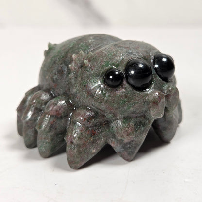 A small carved spider figurine made from polished green malachite with black bead eyes, shown on a light gray surface.