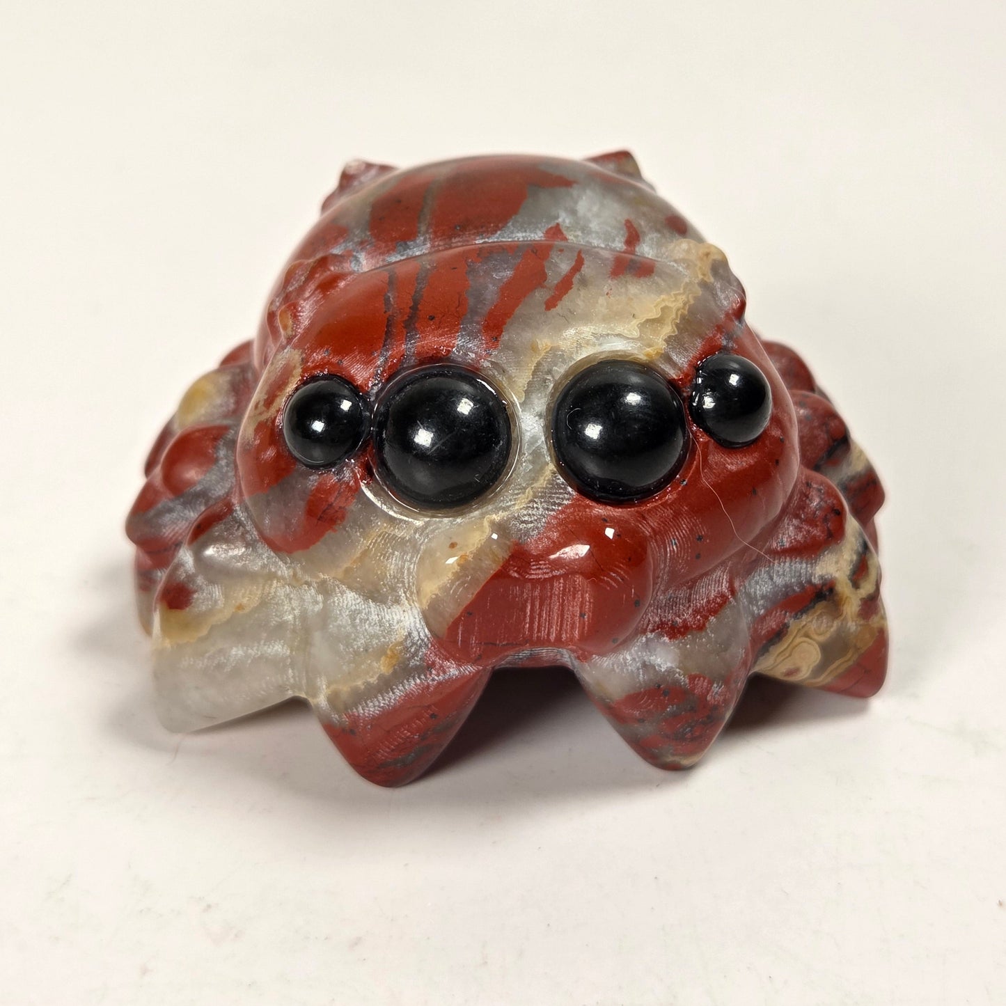 A small decorative spider figurine carved from red and gray agate with black bead eyes, shown on a plain white surface.