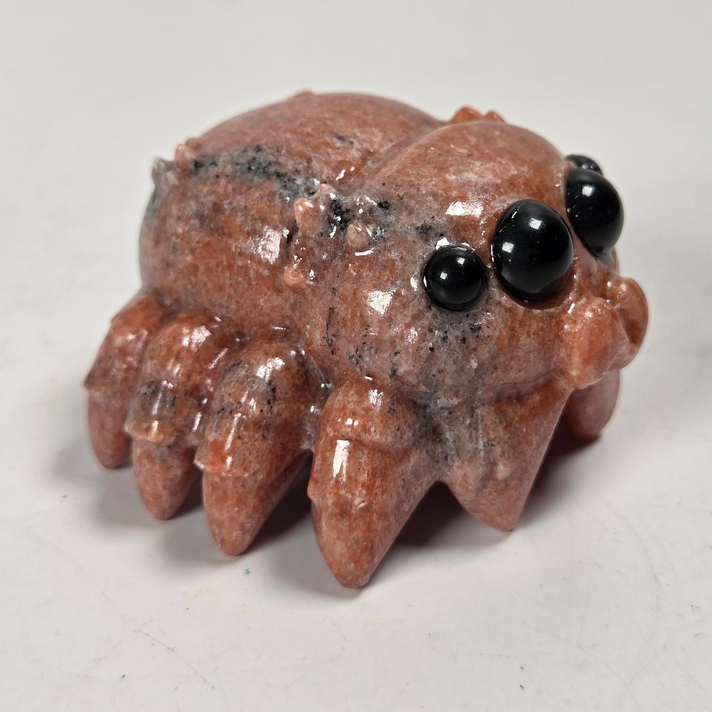 A small carved spider figurine made from polished reddish-brown stone with black gemstone eyes, photographed on a plain light background.