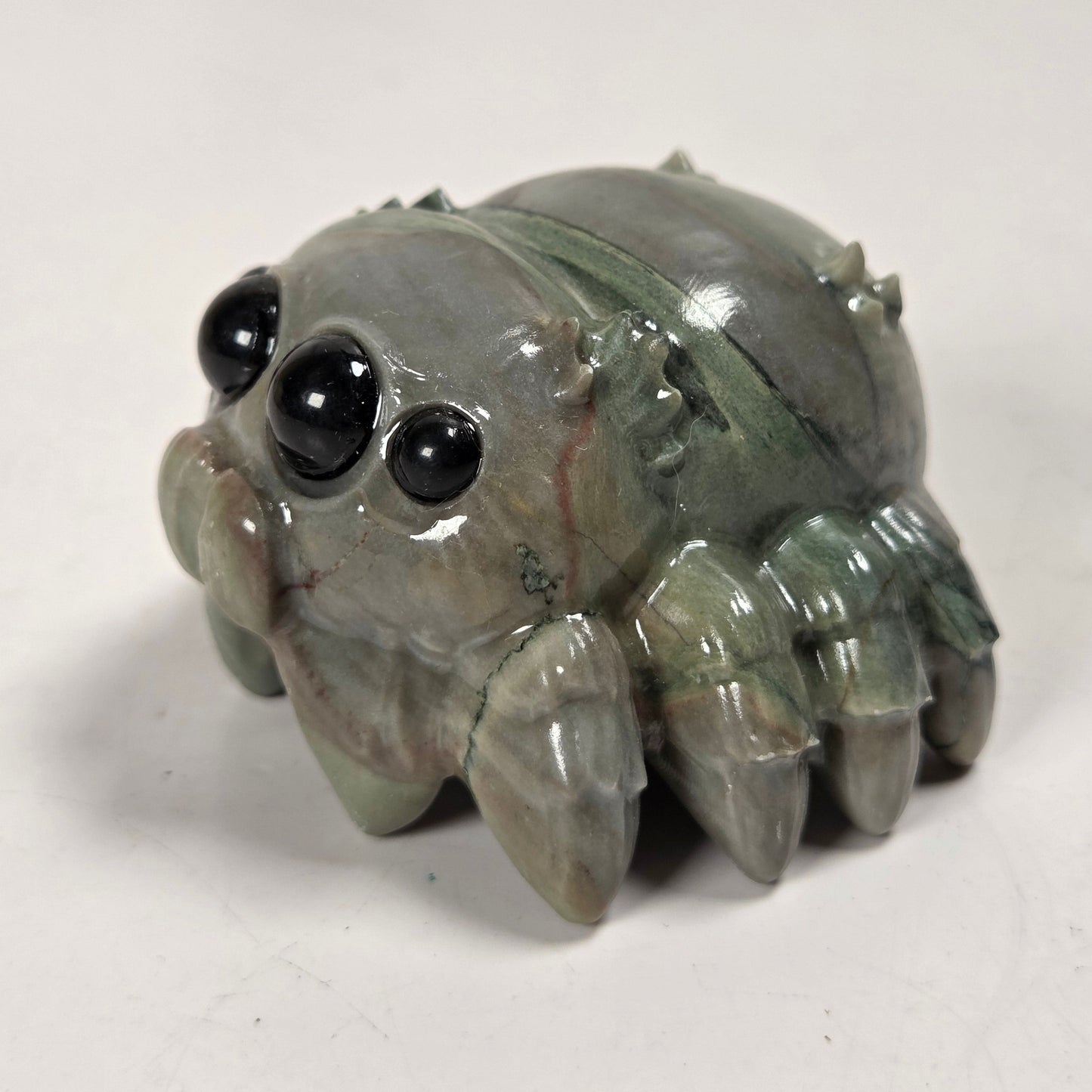 A small carved spider figurine made from polished gray-green stone with large black bead eyes, photographed on a plain light background.