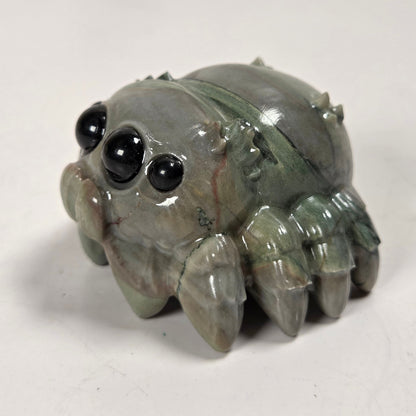 A small carved spider figurine made from polished gray-green stone with large black bead eyes, photographed on a plain light background.