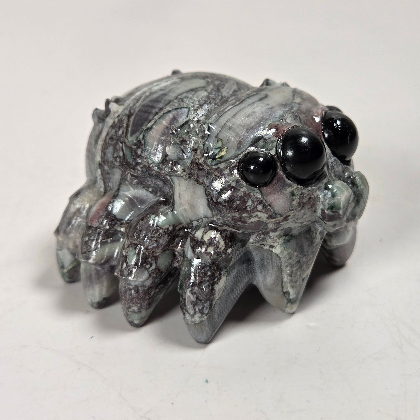 A small spider-shaped decorative object with a glossy, marbled gray and white surface and large black bead eyes, photographed on a plain light background.