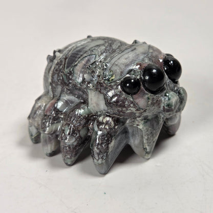 A small spider-shaped decorative object with a glossy, marbled gray and white surface and large black bead eyes, photographed on a plain light background.