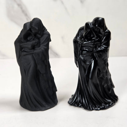 Two black hooded figurines in long flowing robes, standing side by side on a white surface against a light marble-look background.