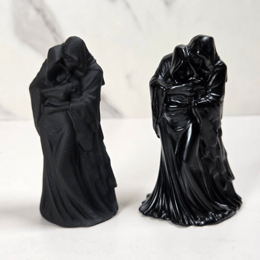 Two black hooded figurines in long flowing robes, standing side by side on a white surface against a light marble-look background.