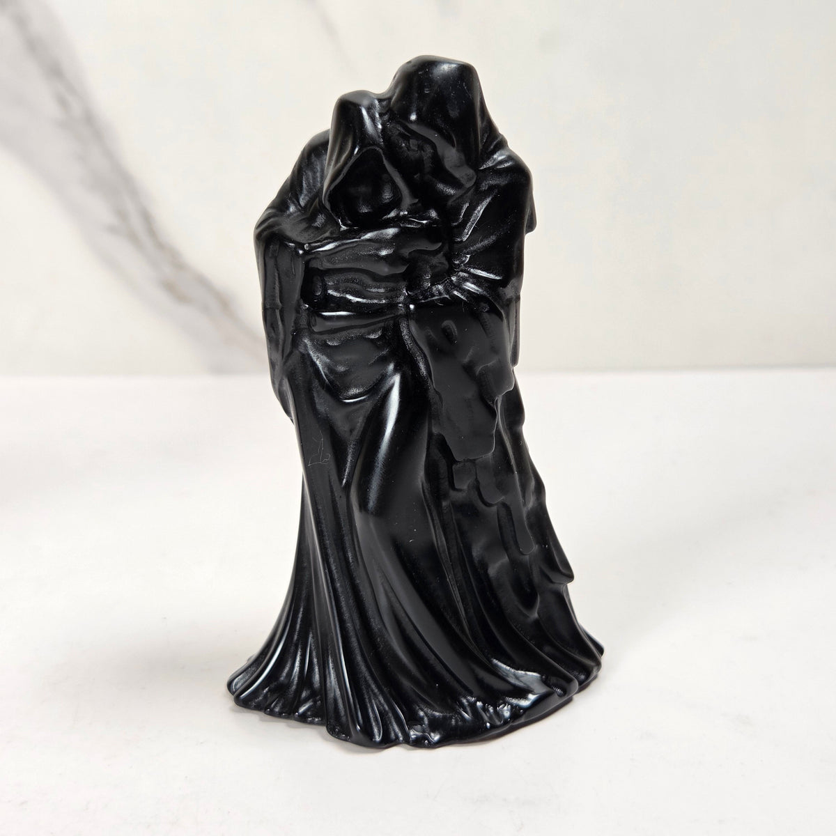 A glossy black figurine of a cloaked, hooded figure with draped robes, standing on a white surface against a light gray marble-patterned background.