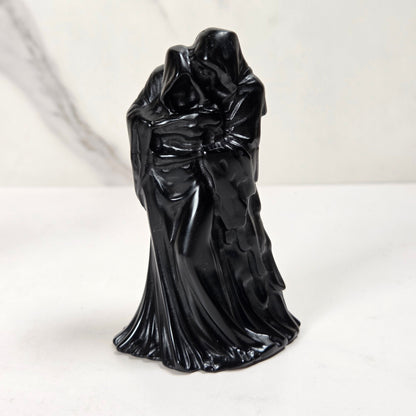 A glossy black figurine of a cloaked, hooded figure with draped robes, standing on a white surface against a light gray marble-patterned background.