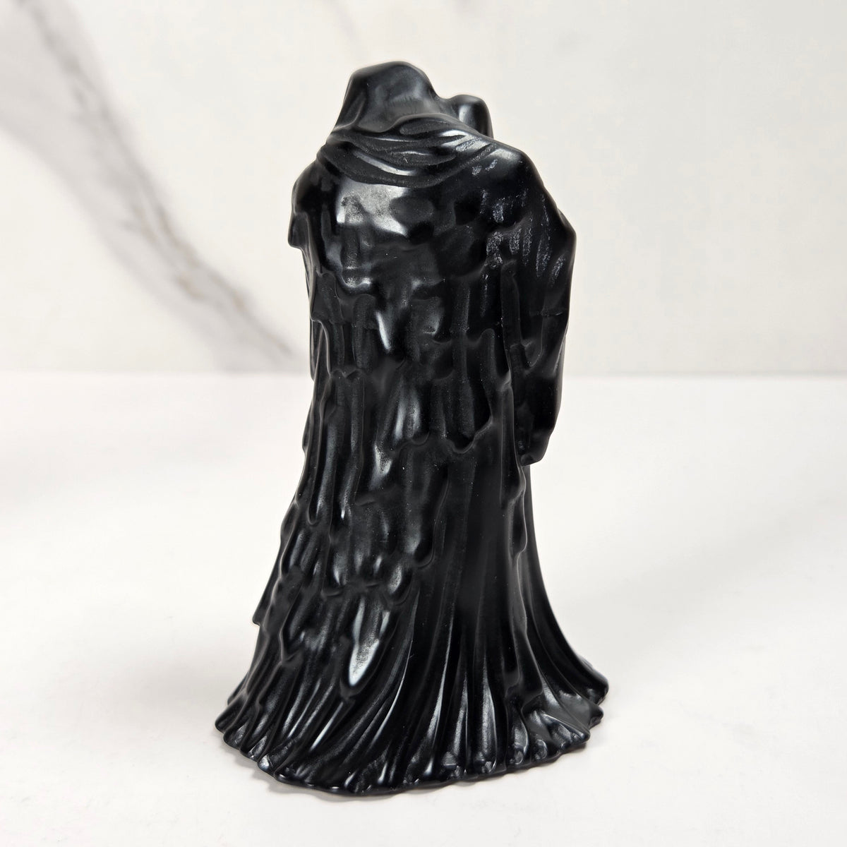 A black hooded ghostly figure figurine with textured, dripping details, standing on a white surface against a light gray marble-patterned background.