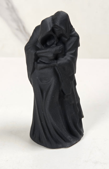 A small black figurine of a draped, hooded robed figure standing on a white surface against a light gray and white marble-patterned background.