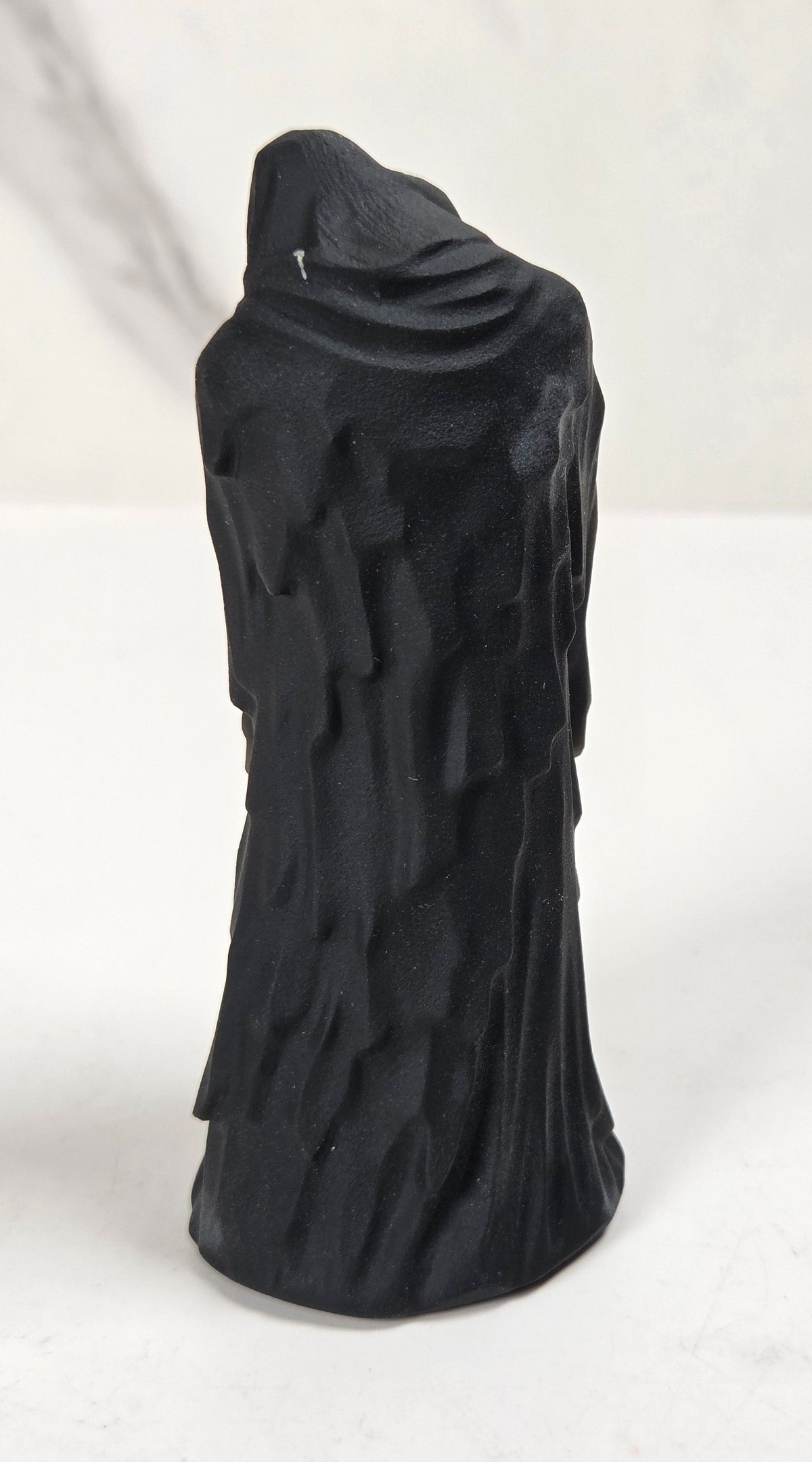 A black textured sculpture of a hooded figure, resembling a grim reaper or raven, standing on a white surface against a light gray marble-patterned background.