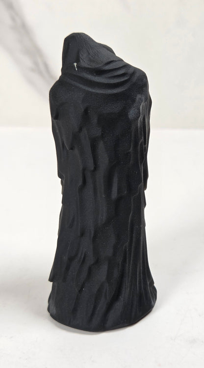 A black textured sculpture of a hooded figure, resembling a grim reaper or raven, standing on a white surface against a light gray marble-patterned background.