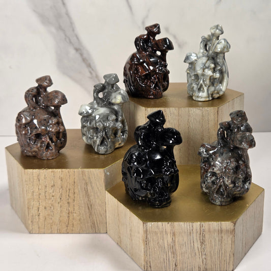 Five small sculptural figurines displayed on stacked hexagonal wooden blocks against a light marble-look background. The figurines appear in glossy dark brown, metallic silver, black, and gray finishes.