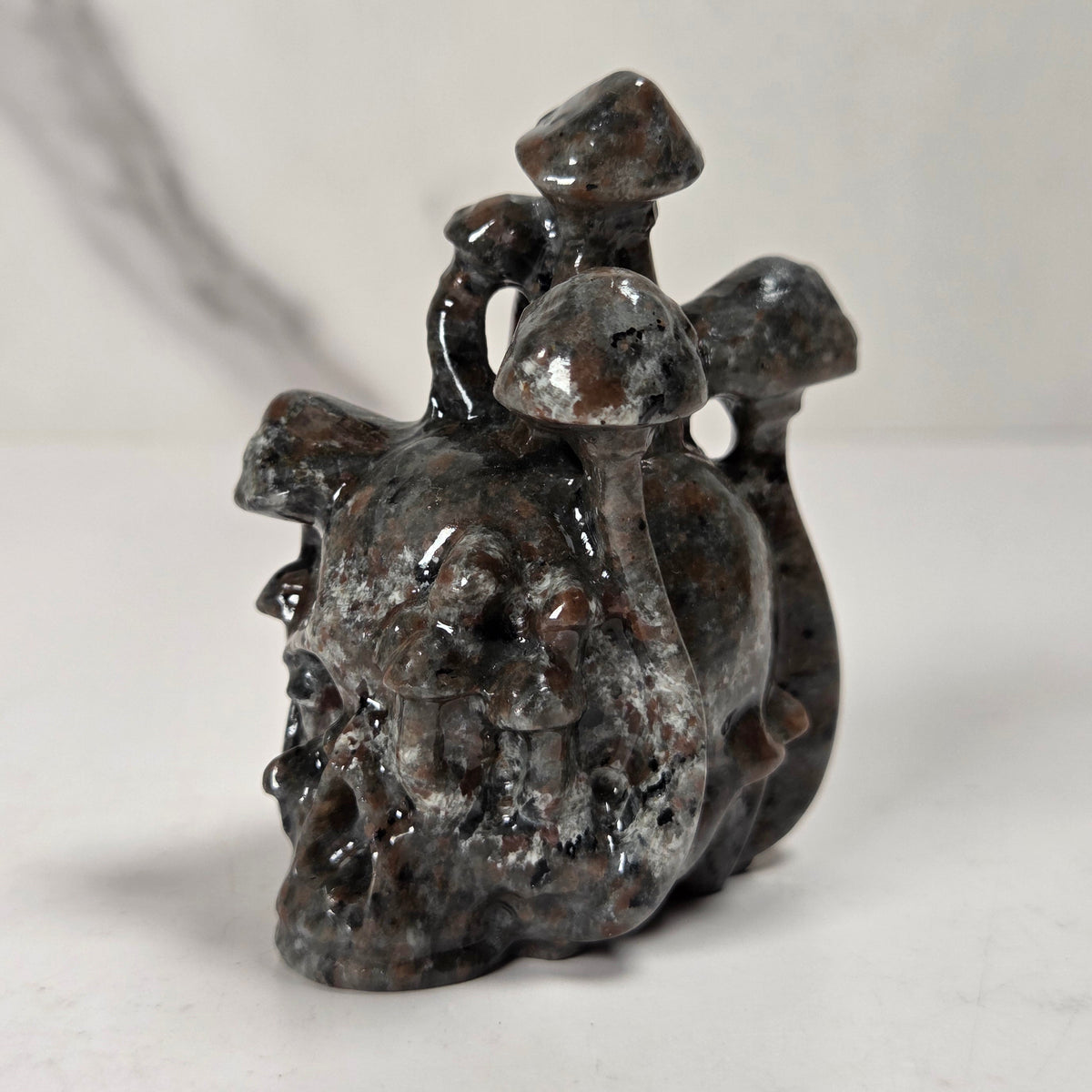 A small carved stone figurine depicting a skull-like form with three mushroom shapes on top, photographed on a light neutral surface with a softly blurred background.