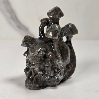 A small carved stone figurine depicting a skull-like form with three mushroom shapes on top, photographed on a light neutral surface with a softly blurred background.