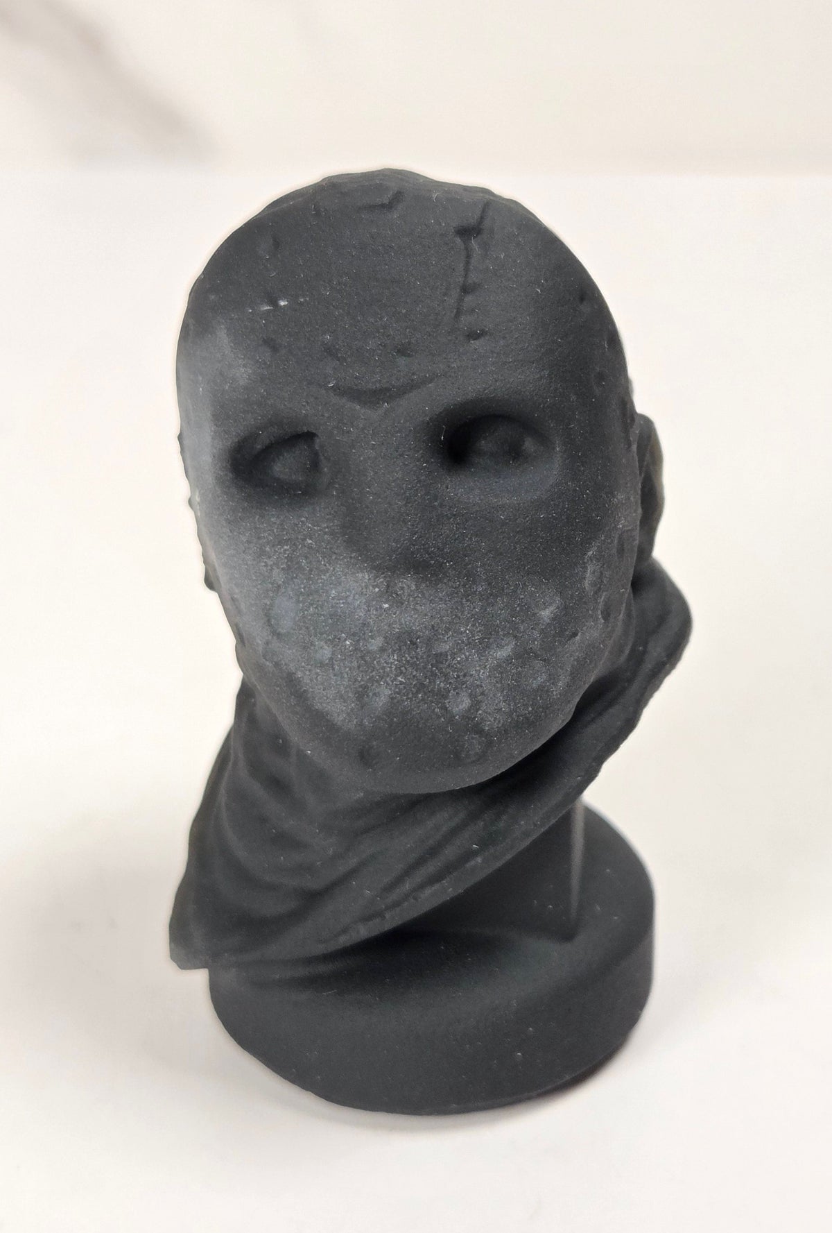 A matte black sculptural bust of a faceless figure with hollow eyes, standing on a round base against a light off-white background.