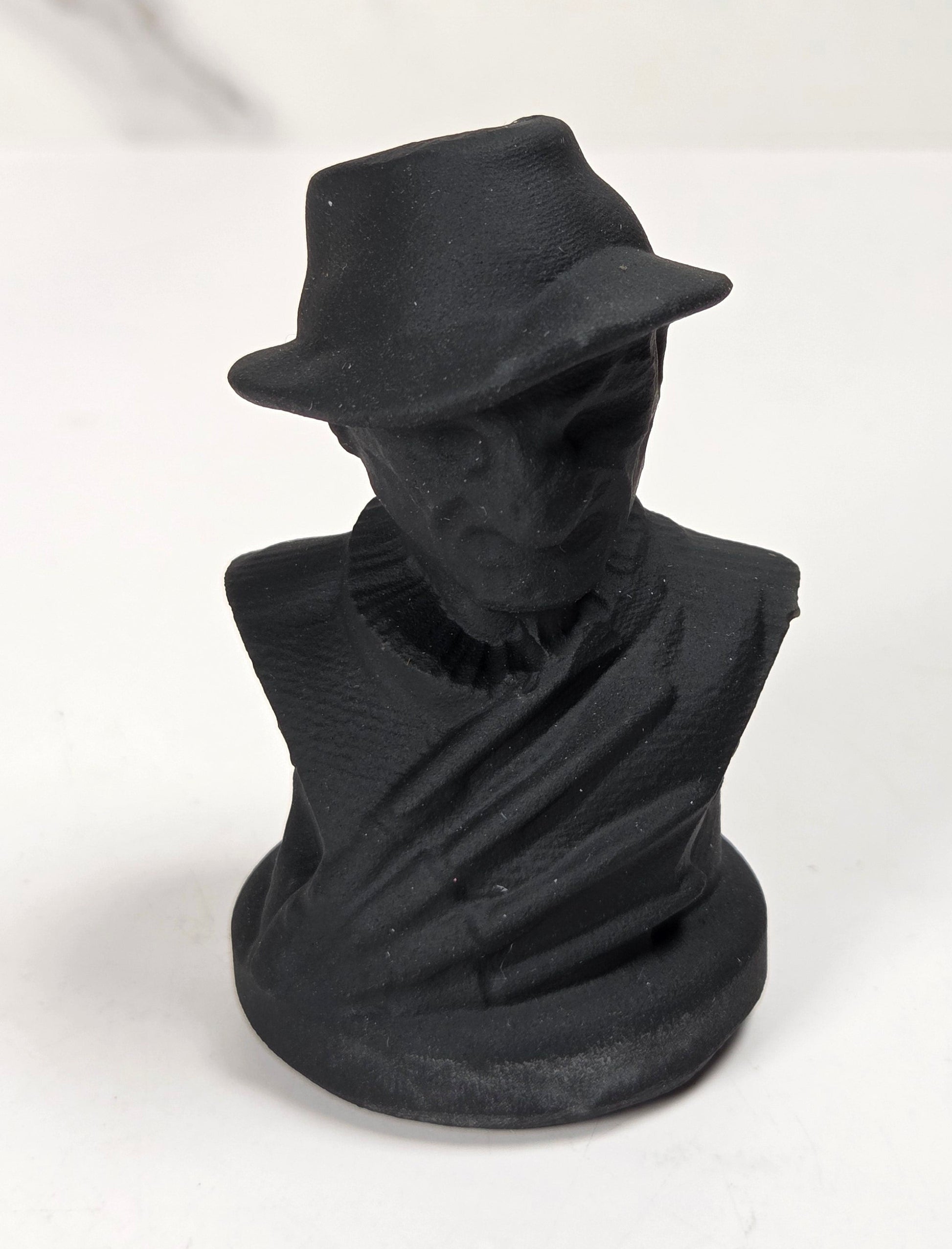 A small matte black sculpture of a man's bust wearing a fedora hat, set against a light gray and white marble-patterned background.