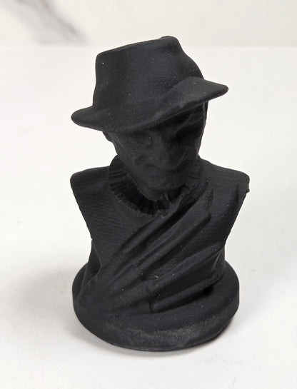 A small matte black sculpture of a man's bust wearing a fedora hat, set against a light gray and white marble-patterned background.