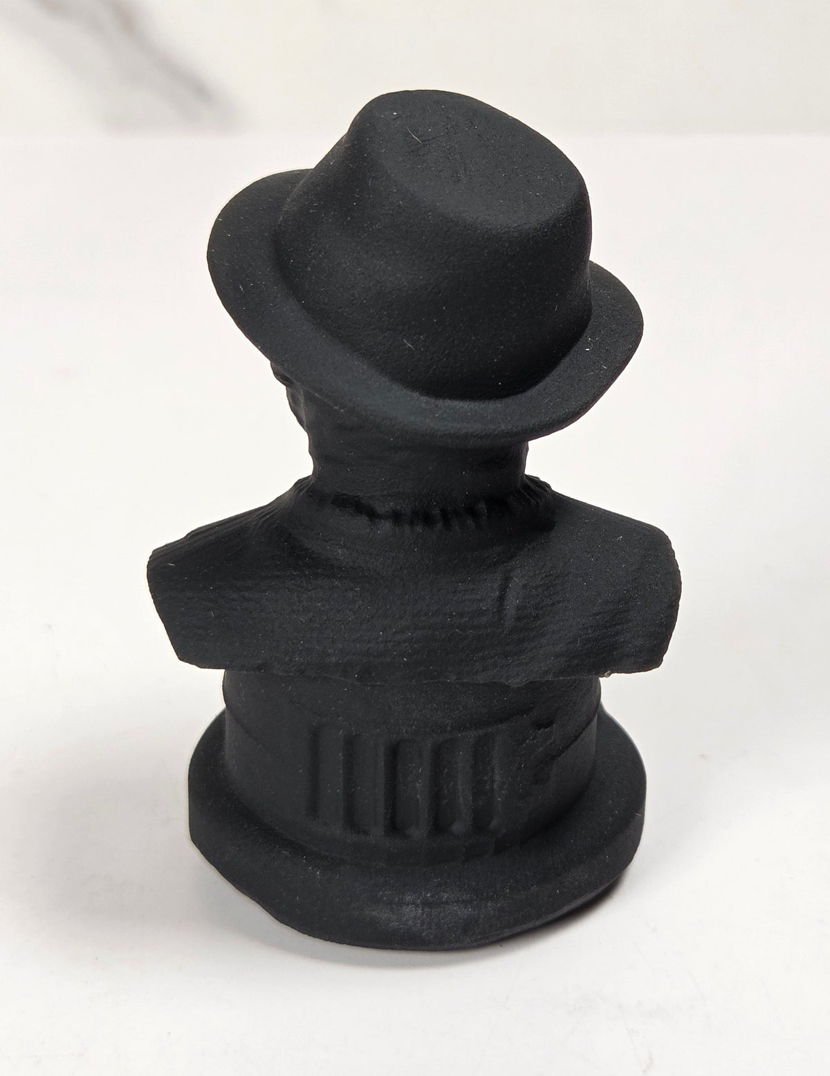 A small matte black figurine of a man's bust wearing a bowler hat, photographed on a white surface with a light gray marble-look background.