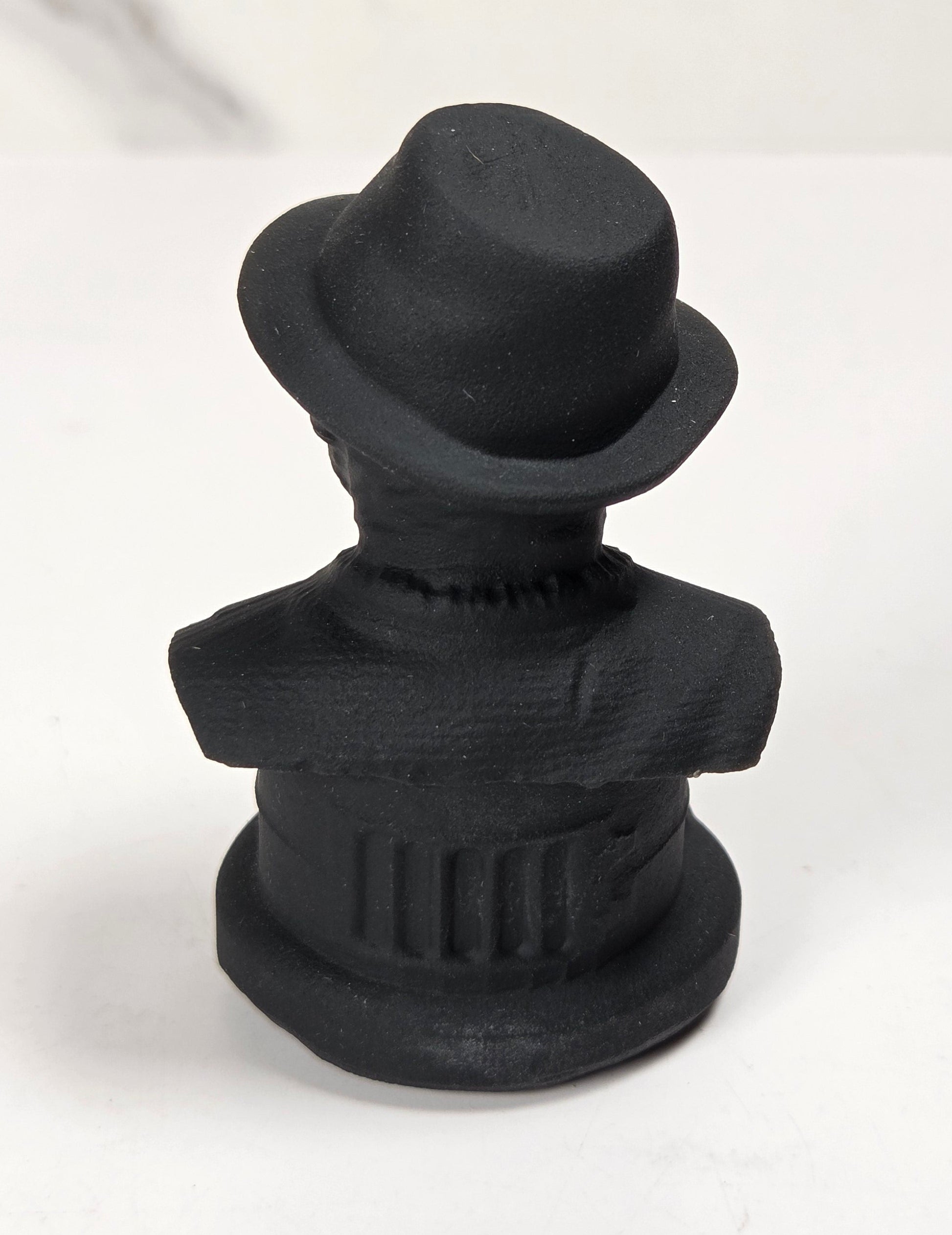 A small matte black figurine of a man's bust wearing a bowler hat, photographed on a white surface with a light gray marble-look background.