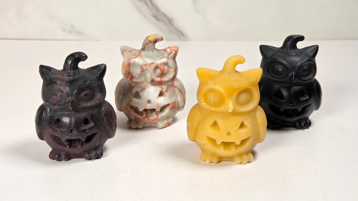Four small owl-shaped jack-o'-lantern candles arranged in a row on a white surface with a light gray marble background. The candles come in four colors: black, white with orange marbling, yellow, and black.