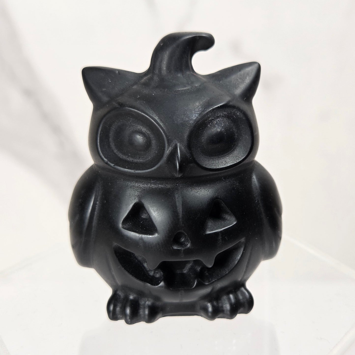 A small black owl-shaped jack-o'-lantern figurine sitting on a clear acrylic display block against a light marble-patterned background.