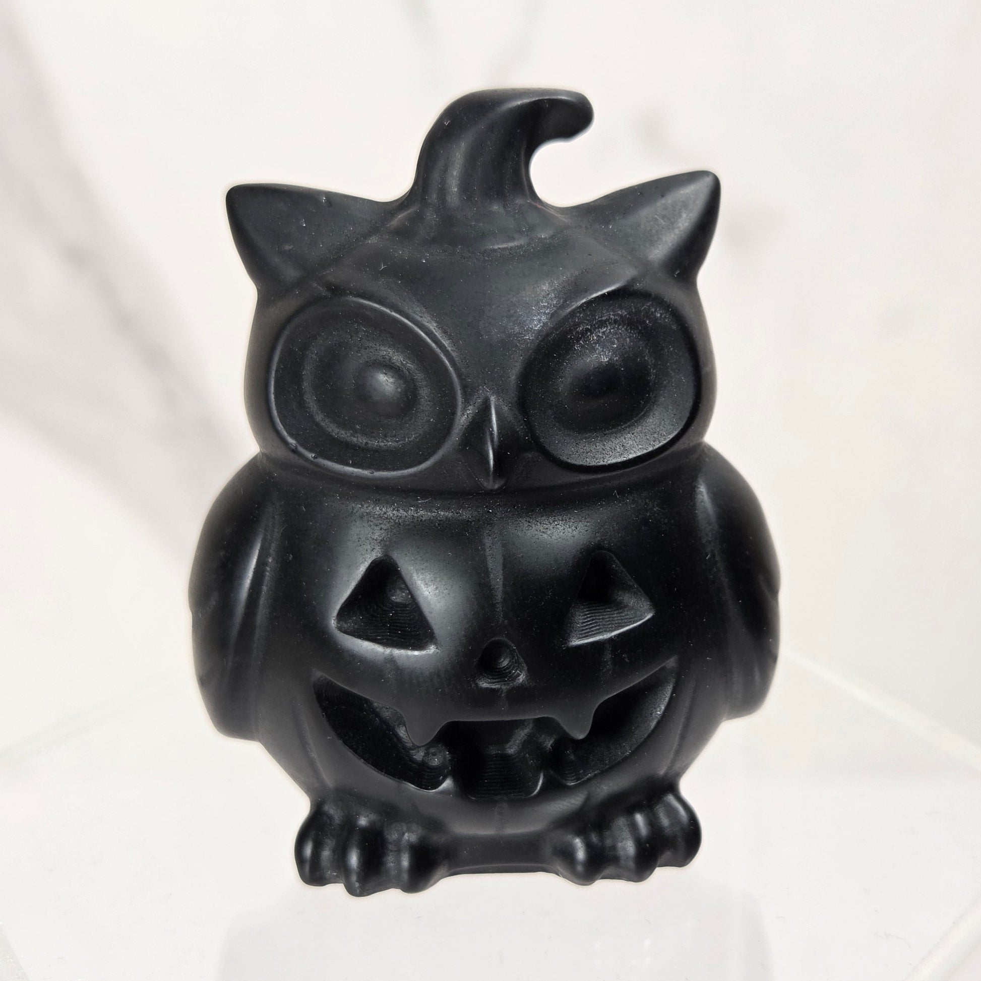 A small black owl-shaped jack-o'-lantern figurine sitting on a clear acrylic display block against a light marble-patterned background.
