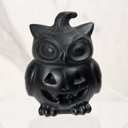 A small black owl-shaped jack-o'-lantern figurine sitting on a clear acrylic display block against a light marble-patterned background.