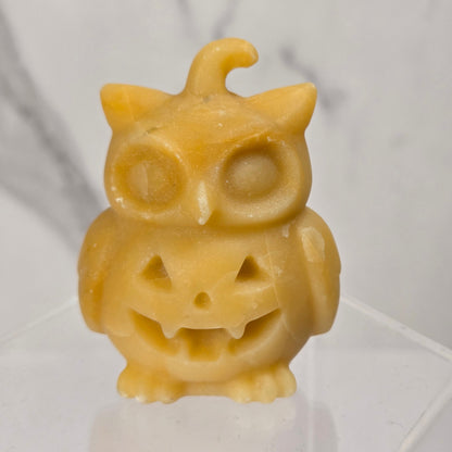 A small yellow owl-shaped candle with a carved jack-o'-lantern face and a curled top tail, displayed on a clear acrylic stand against a light gray marble-patterned background.