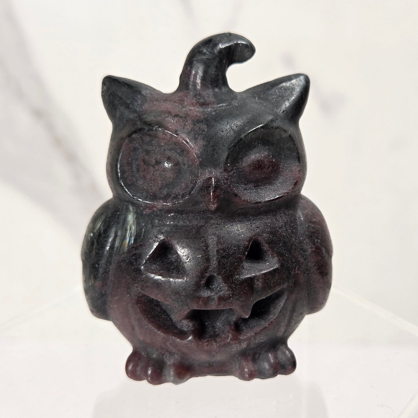 A small carved decorative owl figurine with a jack-o'-lantern face and a witch hat, shown standing on a clear acrylic display block against a light marble-like background.