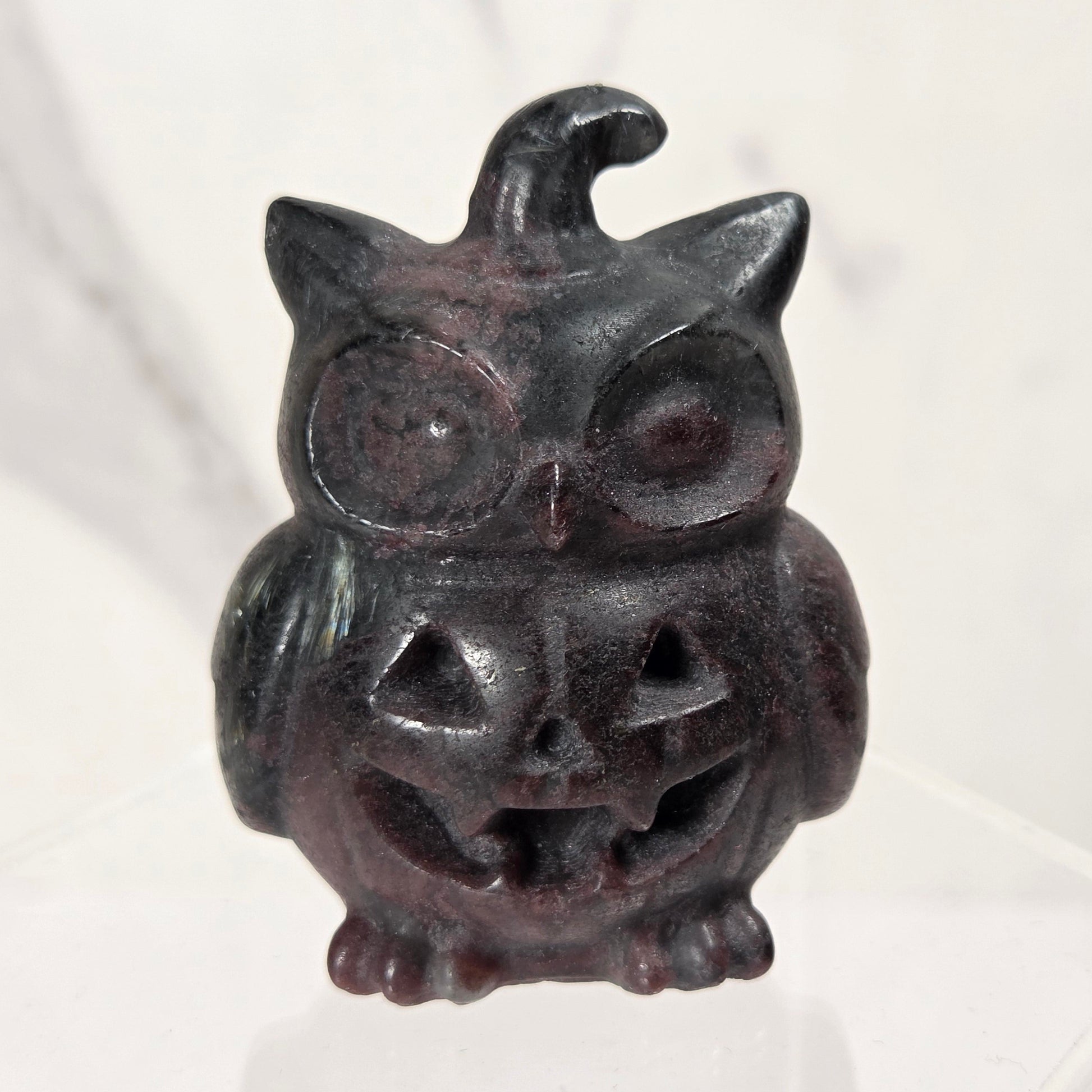 A small carved decorative owl figurine with a jack-o'-lantern face and a witch hat, shown standing on a clear acrylic display block against a light marble-like background.