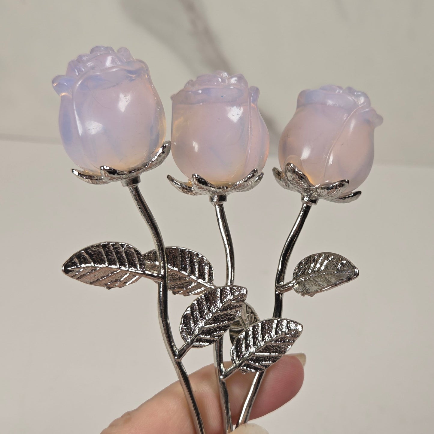 Three rose-shaped brooches with iridescent lavender-pink petals and silver-toned stems and leaves, held in a hand against a light marble-patterned background.