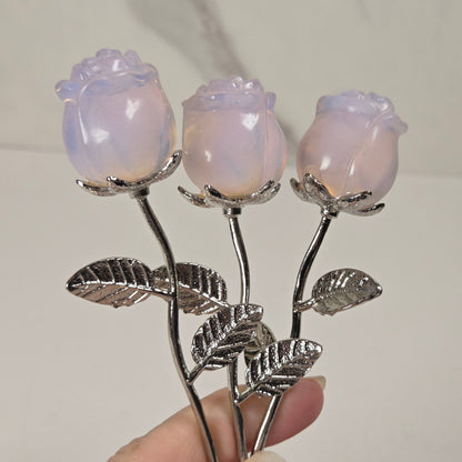 Three rose-shaped brooches with iridescent lavender-pink petals and silver-toned stems and leaves, held in a hand against a light marble-patterned background.