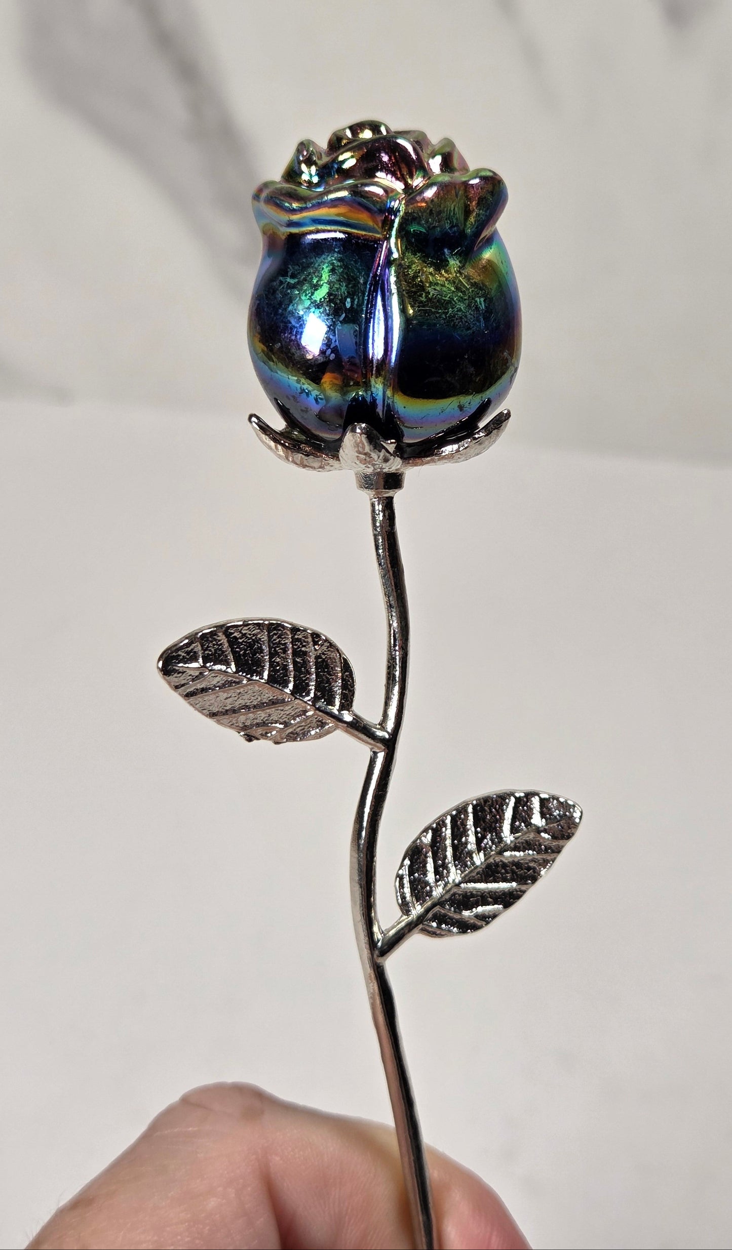 A hand holding a decorative metal rose with a silver stem and leaves, topped with an iridescent blue and green glass rosebud, against a light gray marble-patterned background.