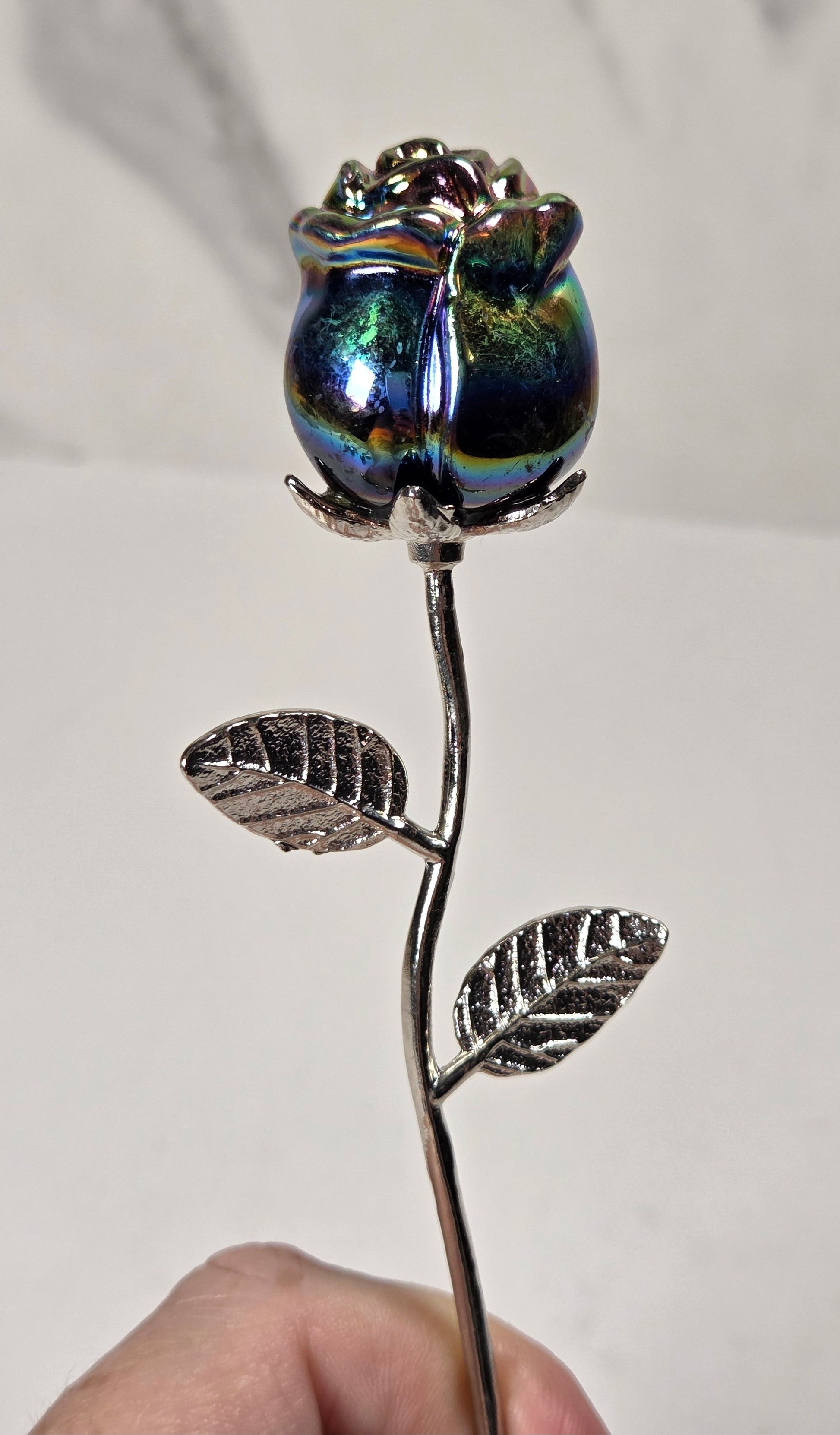 A hand holding a decorative metal rose with a silver stem and leaves, topped with an iridescent blue and green glass rosebud, against a light gray marble-patterned background.