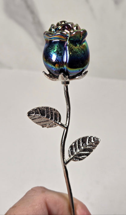 A hand holding a decorative metal rose with a silver stem and leaves, topped with an iridescent blue and green glass rosebud, against a light gray marble-patterned background.