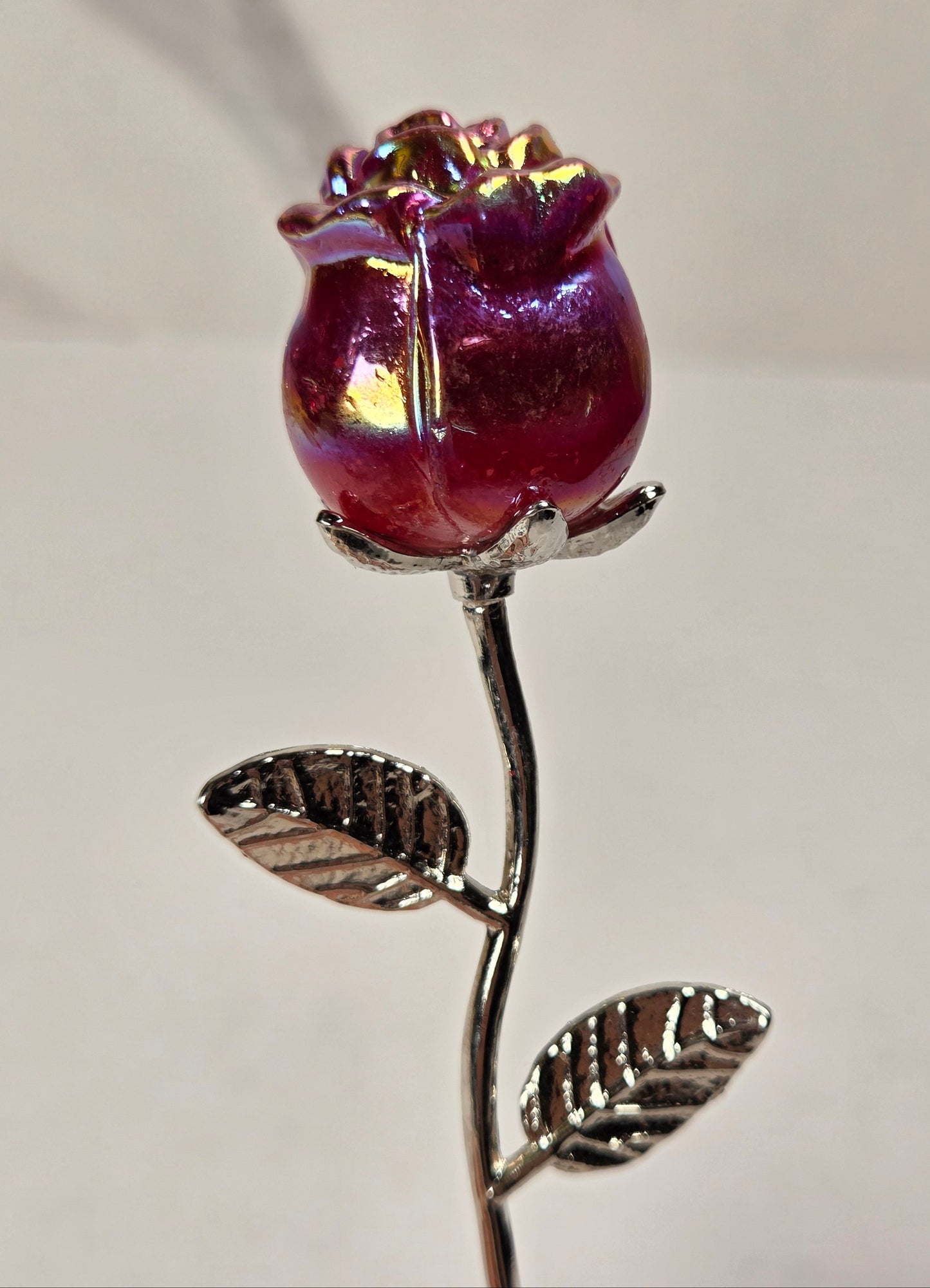 A single rose-shaped decorative object with an iridescent deep pink and purple bloom on a metallic silver stem and leaves, photographed against a plain light background.