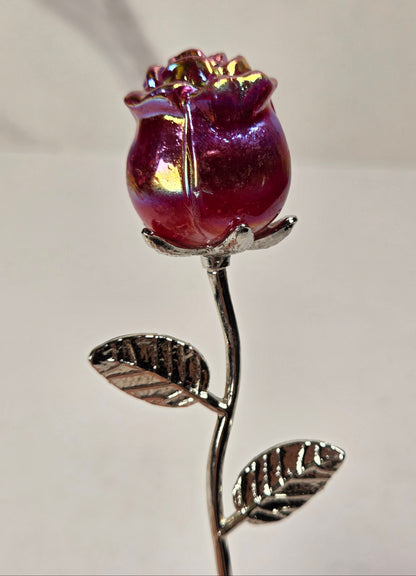 A single rose-shaped decorative object with an iridescent deep pink and purple bloom on a metallic silver stem and leaves, photographed against a plain light background.
