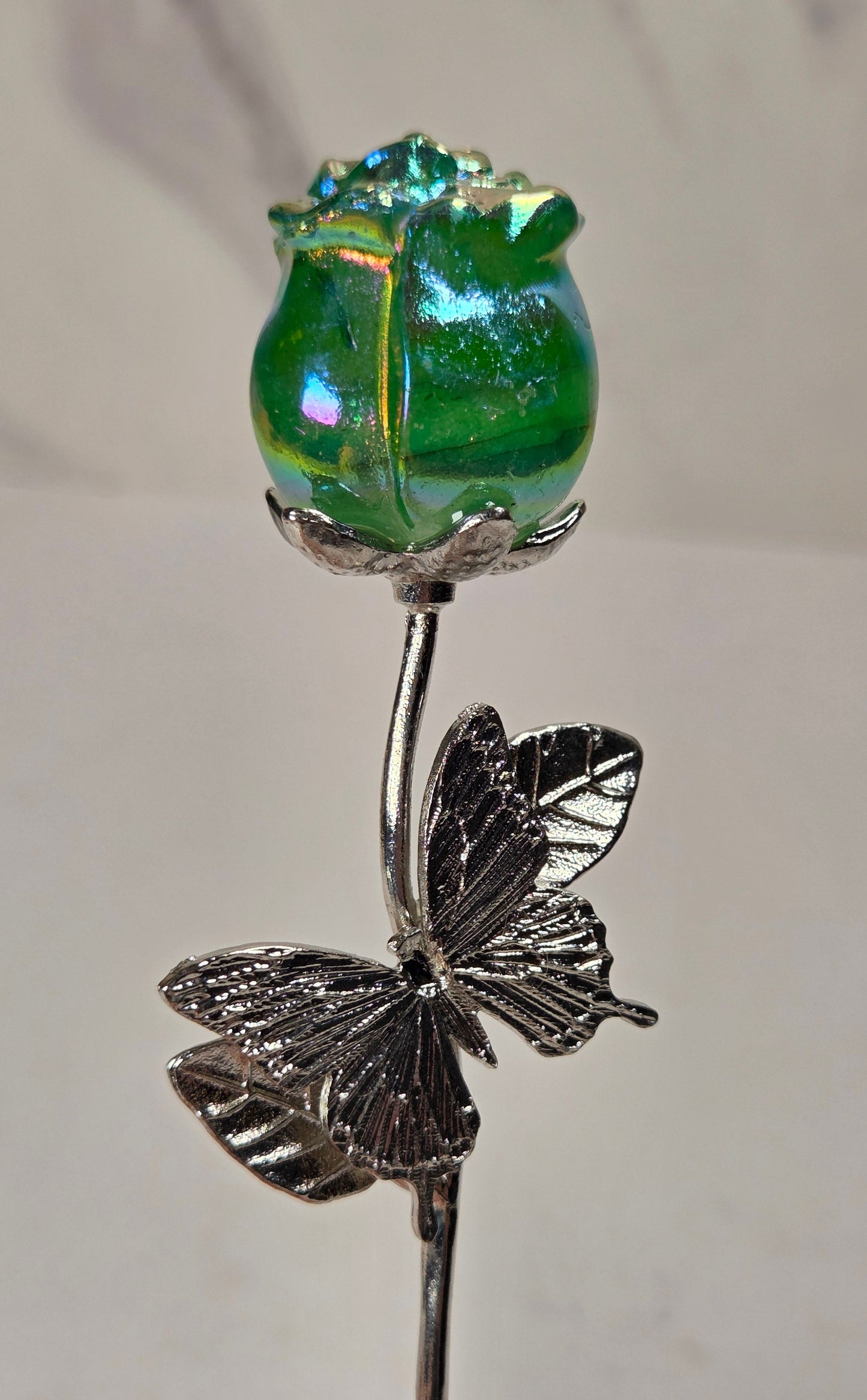 A decorative glass and metal flower pin featuring a green iridescent glass rose bud on a silver-toned stem with a butterfly motif, held against a light neutral background.