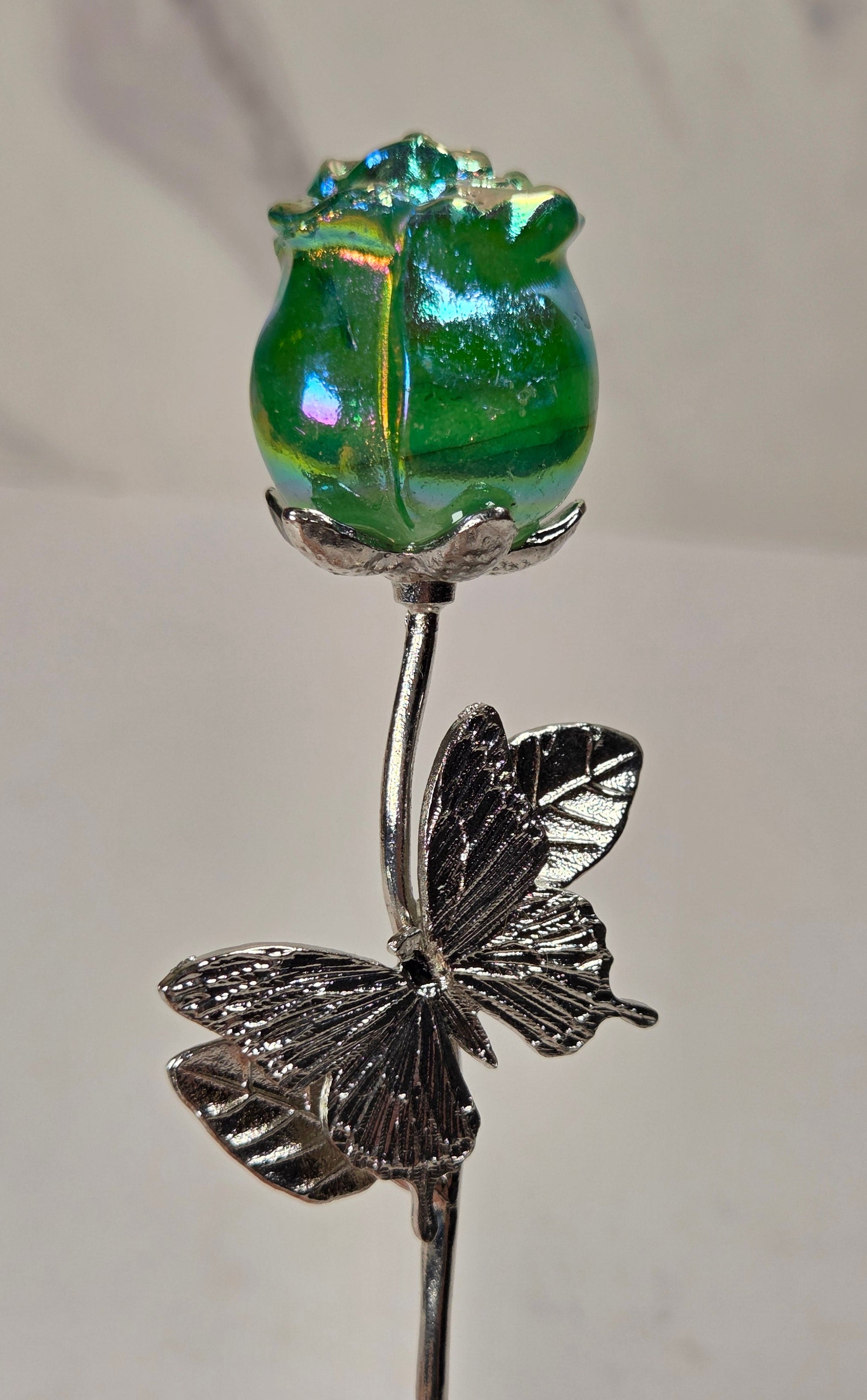 A decorative glass and metal flower pin featuring a green iridescent glass rose bud on a silver-toned stem with a butterfly motif, held against a light neutral background.