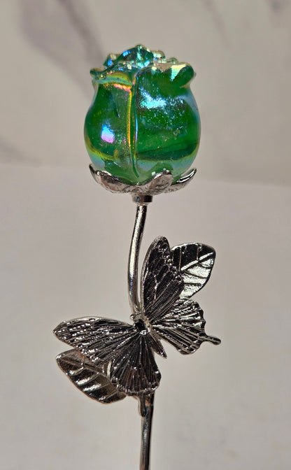 A decorative glass and metal flower pin featuring a green iridescent glass rose bud on a silver-toned stem with a butterfly motif, held against a light neutral background.