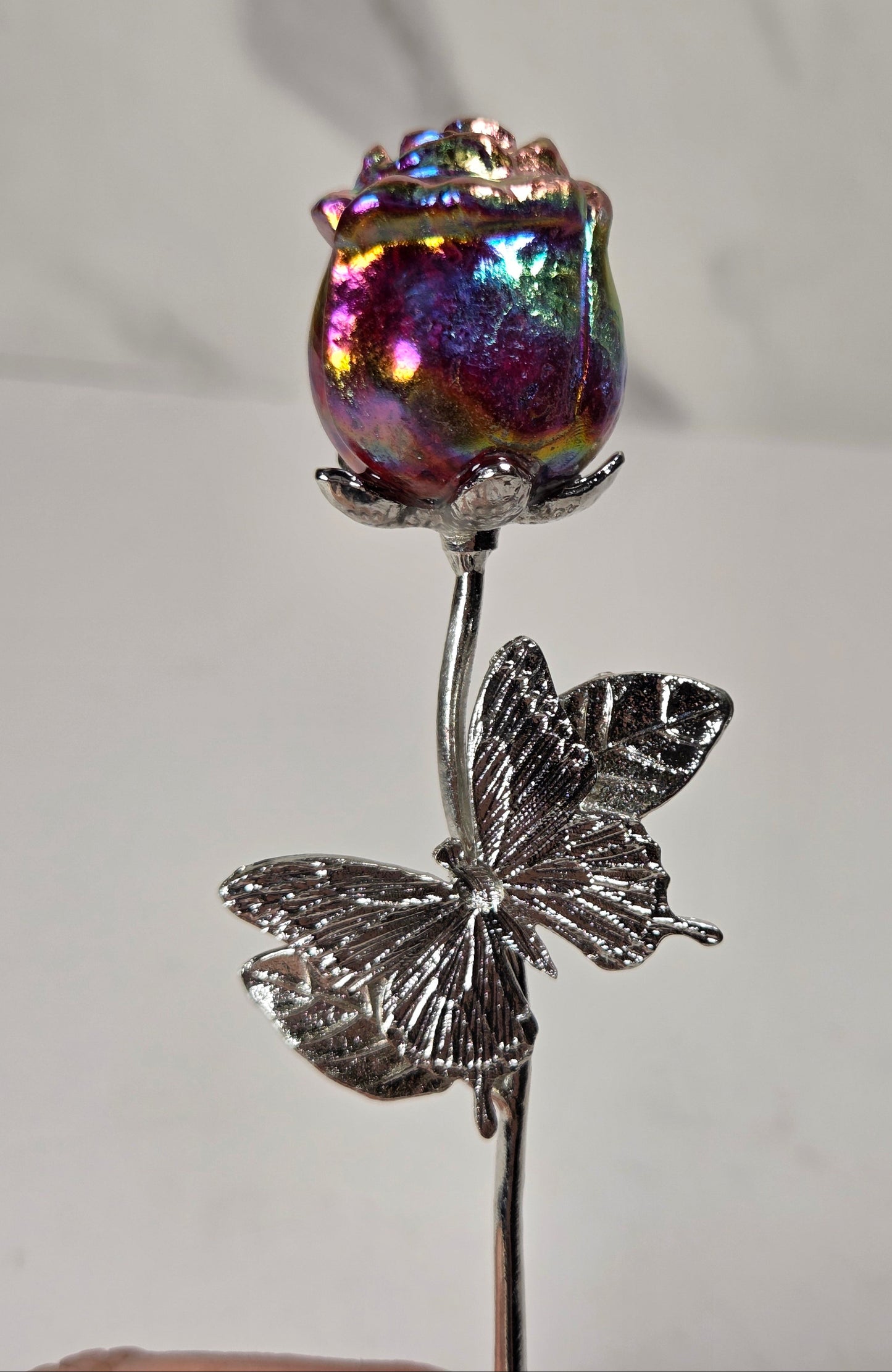 A decorative metal rose pin featuring a silver butterfly stem and a vibrant, iridescent holographic rosebud, held against a light gray background.