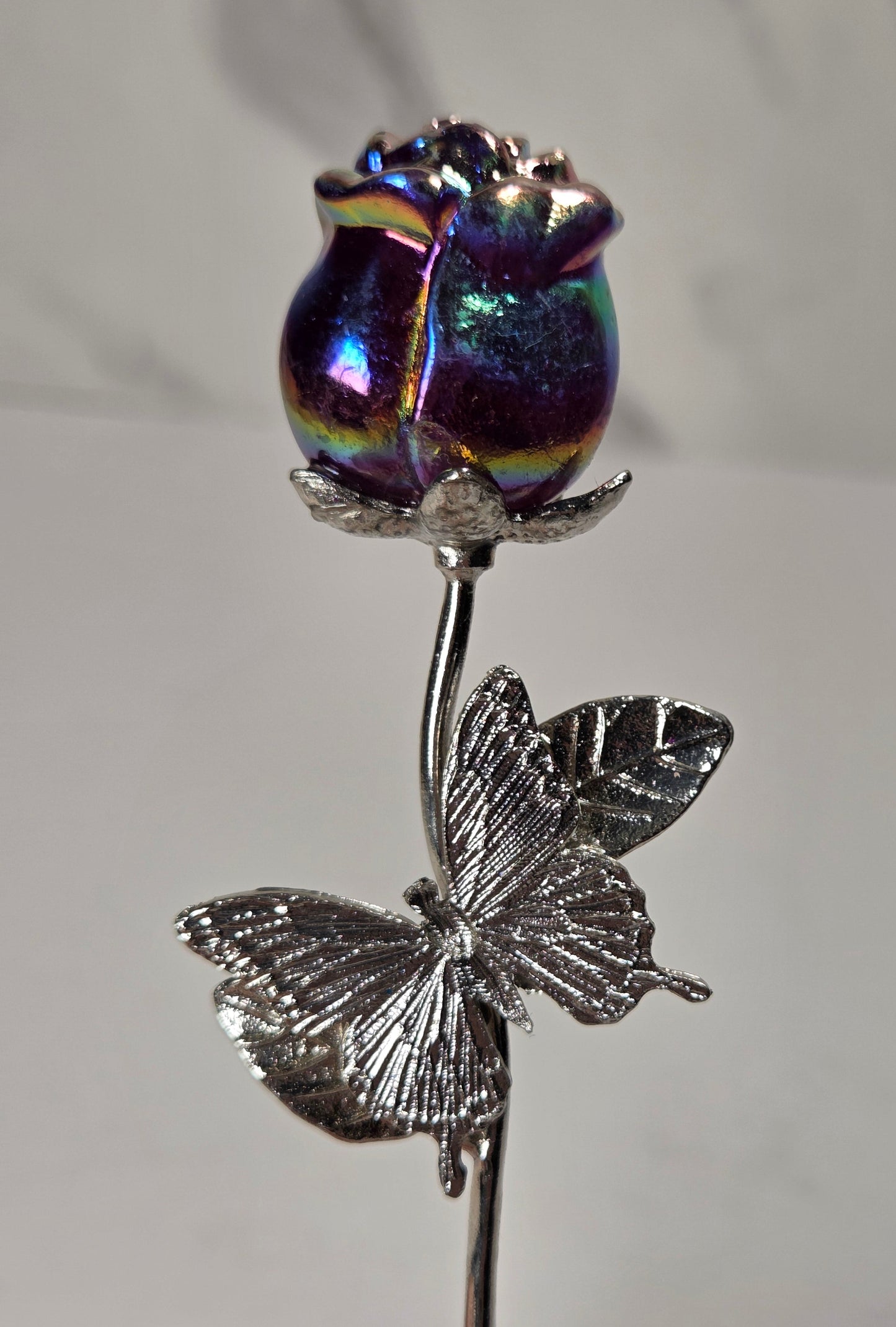 A silver butterfly and stem flower ornament with a highly iridescent, holographic purple rosebud on top, held by a human finger against a neutral gray background.