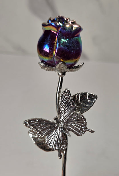 A silver butterfly and stem flower ornament with a highly iridescent, holographic purple rosebud on top, held by a human finger against a neutral gray background.
