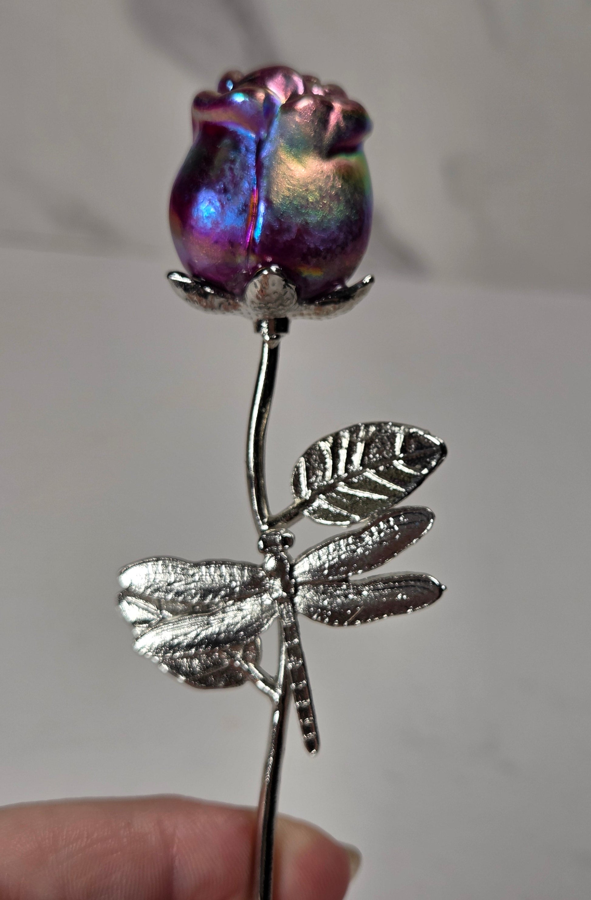 A close-up of a hand holding a decorative rose-shaped object with an iridescent purple and blue glass rose bud on a silver stem and dragonfly motif.