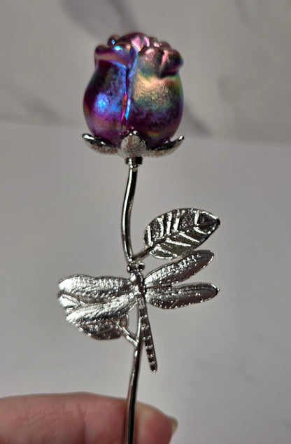 A close-up of a hand holding a decorative rose-shaped object with an iridescent purple and blue glass rose bud on a silver stem and dragonfly motif.