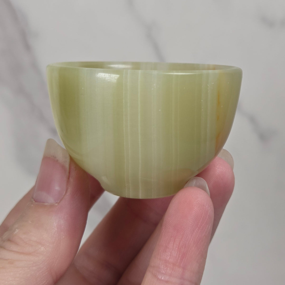 A small, rounded cup made of pale yellow-green stone, held between fingers against a light marble-patterned background.